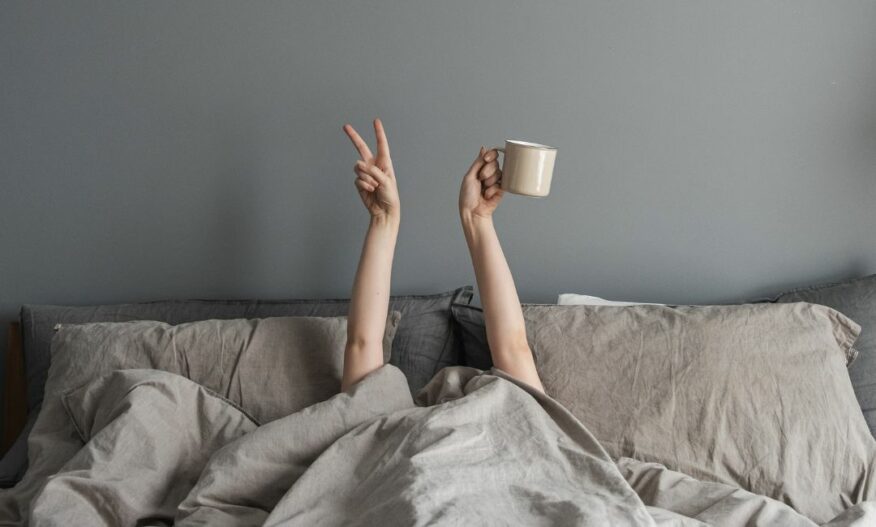 Dame ligger under dyne i seng, hendene opp i lufte med kaffekopp. IMG