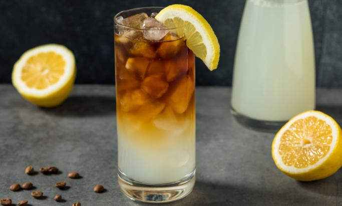 Et glass med kaffelemonade står på en grå kjøkkenbenk, ved siden av oppskjært sitron og kaffebønner